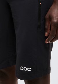 Κοντινό πλάνο ενός ατόμου που φοράει μαύρα σορτς με το λογότυπο POC, με τσέπη με φερμουάρ, και το χέρι ακουμπισμένο κοντά στη ραφή του πλαινού.