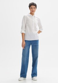 Camicetta bianca in lino con colletto, maniche arrotolate e taschino sul petto, abbinata a jeans a gamba larga di colore blu chiaro e scarpe da ginnastica bianche.