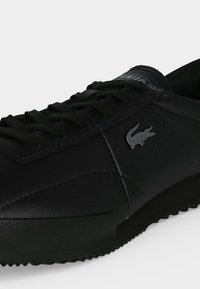 Μαύρο χαμηλό sneakers με μαύρα κορδόνια, υφασμένο πάτο και ασημί λογότυπο κροκόδειλου στη πλαϊνή πλευρά.