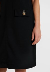 Černé šaty s kapsou a kulatým knoflíkem. Materiál vypadá hladce a má upravený tvar; ruka je umístěna blízko detailu kapsy.