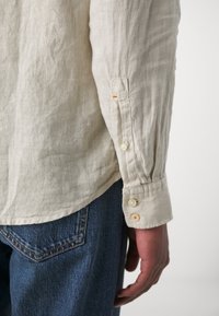 Camisa de lino beige claro con puños abotonados, que presenta un sutil hilo naranja. Combinada con vaqueros de denim azul oscuro.