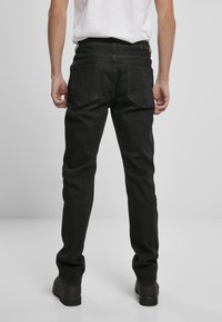 Crne traperice s ravnim nogavicama, s dva stražnja džepa i oznakom na remenu. Materijal izgleda čvrsto s glatkom teksturom.