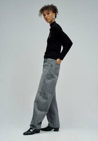 Młoda kobieta z kręconymi włosami, ubrana w czarny golf, szare szerokie spodnie dżinsowe i czarne botki za kostkę, stojąca w profilu z jedną ręką w kieszeni.