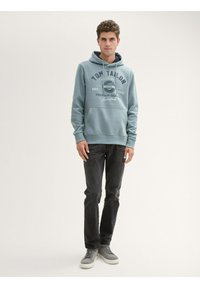 Felpa blu chiaro con cappuccio, dotata di tasca frontale e logo stampato. Abbinata a jeans scuri e scarpe da ginnastica grigie, in piedi contro uno sfondo neutro.