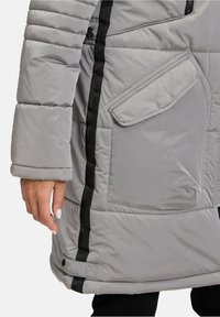 Doudoune grise avec une poche latérale, dotée d'une texture côtelée, de sangles noires décoratives et d'une coupe décontractée. Détail du bas ajustable visible.