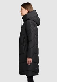 Svart vadderad jacka med hög, justerbar huva, lång modell, diagonalt quiltat mönster och fickor med dragkedja i sidorna. Har en logotypdetalj.