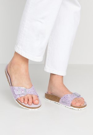 Person iført hvide bukser og lyse lilla sliders med korksål, sølvspænde og tekstureret overdel, stående på et hvidt gulv.