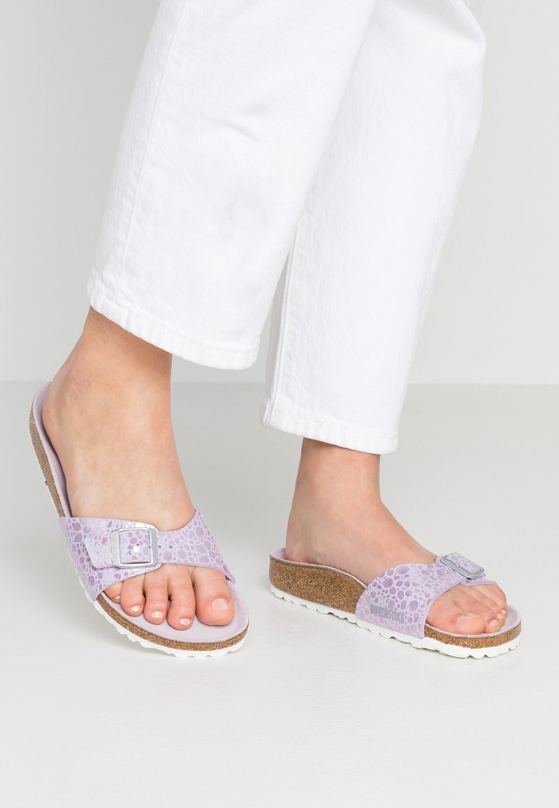 Personne portant un pantalon blanc et des sandales à semelle en liège violet clair avec une boucle argentée et une tige texturée, debout sur un sol blanc.