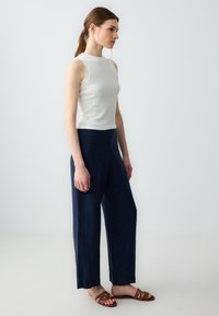 Witte mouwloze top met een gestructureerd patroon, gecombineerd met marineblauwe wijde broek. Bruine sandaaltjes met banden maken de look compleet. Minimalistisch ontwerp.