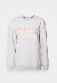 Sudadera gris con cuello redondo, que presenta el texto en tono dorado "TOMMY JEANS", estrellas y un dobladillo acanalado. Ajuste casual, textura suave.