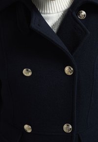 Marineblå uldfrakke med dobbeltbrystede guldknapper, en struktureret krave og en ribbet hvid sweater under.