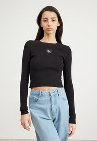 Sort, ribbet langærmet crop top med firkantet udskæring og et lille logo-patch; kombineret med lyseblå høj talje jeans.