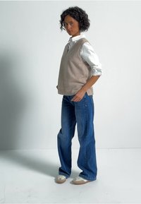 Beige stickad väst över en vit skjorta med knappar, i kombination med avslappnade blå denimjeans och ljusa sneakers.