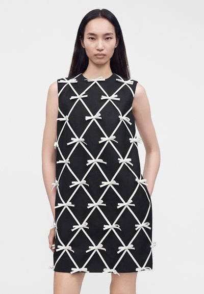 Femme portant une robe noire sans manches avec un motif blanc en croisillons et de petits nœuds blancs à chaque intersection, debout sur un fond blanc.