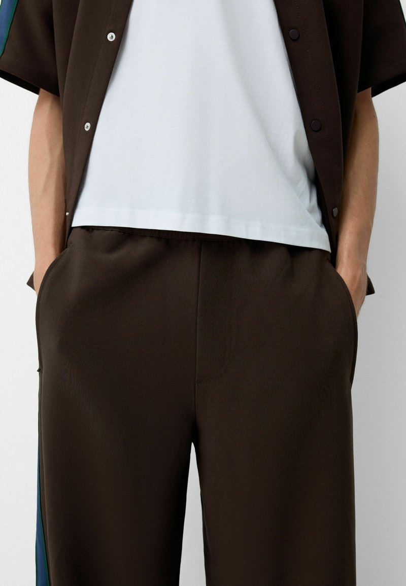 Un pantalon marron ample avec des poches latérales, une texture lisse et une ceinture élastique, associé à une chemise bleu clair sous une veste sombre.