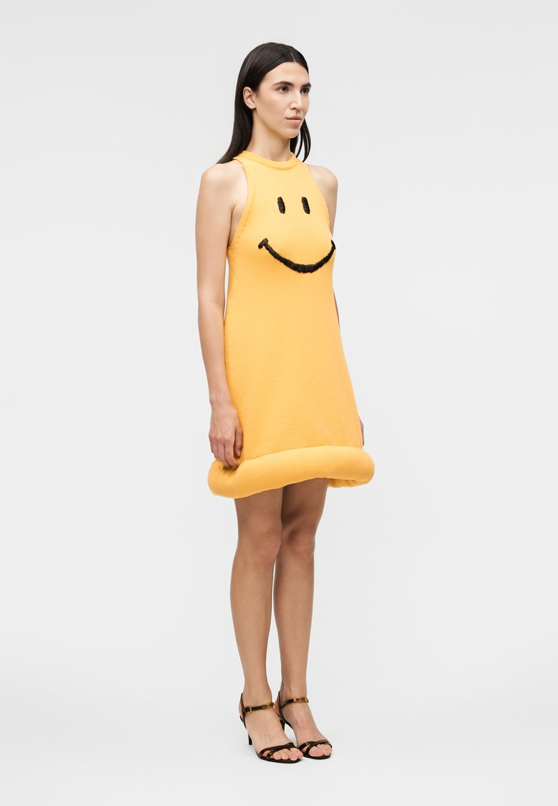 Woman wearing sleeveless yellow dress with black smiley face design and rolled-up hem, paired with black open-toe heels.