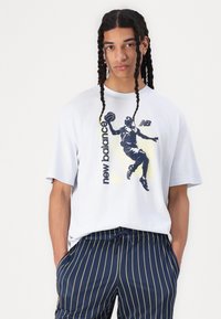 Balts īsām piedurknēm T-krekls ar melnu basketbola grafiku un tekstu "new balance". Saskaņots ar tumši ziliem svītrainiem šortiem.