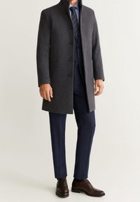 Abrigo de lana gris oscuro con un alto cuello, diseño de botones en la parte delantera, usado sobre un traje azul marino con pantalones a cuadros y zapatos de cuero marrón.
