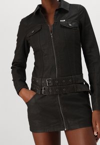 Svart denimjacka klänning med dragkedja, krage, två bröstfickor och bälte i midjan med runda utskärningar. Detaljerad sömnad synlig.