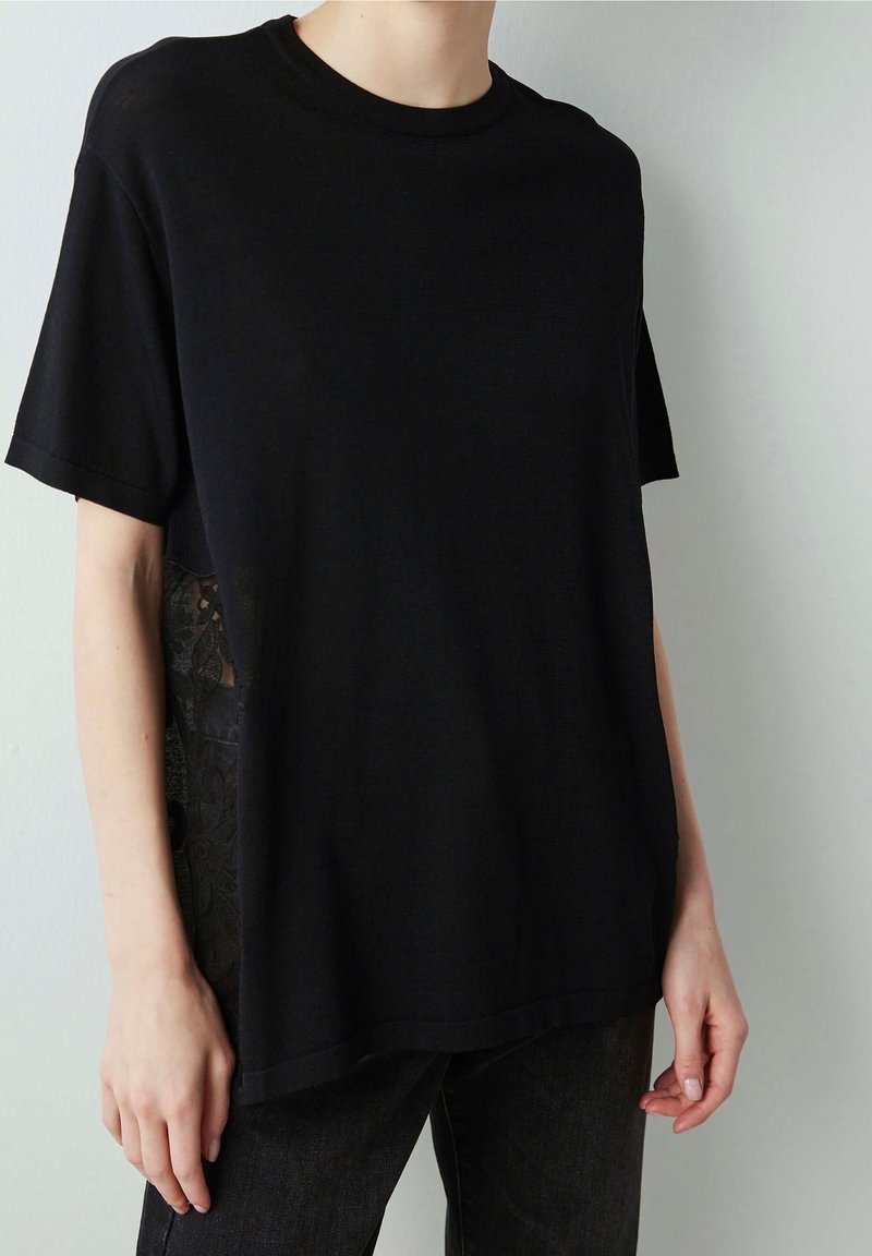 Personne portant une chemise noire ample à manches courtes avec des détails subtils en dentelle sur les côtés et un pantalon foncé sur un fond clair.