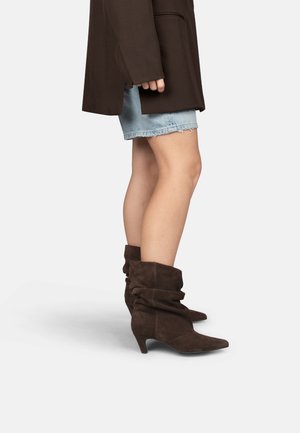 Persoon draagt lichtblauwe spijkershorts, een donkerbruine jas en losse donkerbruine suède enkellaarzen met lage hakken.