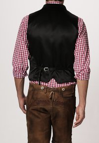 Schwarze Satinveste mit einem tief ausgeschnittenen Rücken und verstellbarem Träger, getragen über einem rot-weiß karierten Hemd, kombiniert mit braunen Lederhosen mit dekorativer Naht.