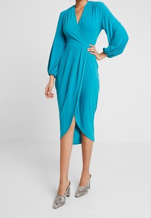 Vrouw in een teal wikkeljurk met lange pofmouwen en een voorsplit, gecombineerd met hoge hakken met slangenhuidpatroon.