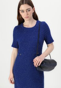 Gebreid jurk in gestructureerd blauw, met korte mouwen, ronde halslijn en twee decoratieve gouden hardwareaccenten. Zwarte handtas met zachte afwerking.