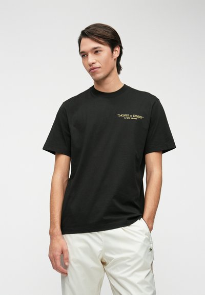 Черна памучна тениска с къси ръкави, с жълт текст "LACOSTE x TENNIS" отпред. Моделът носи светли панталони.