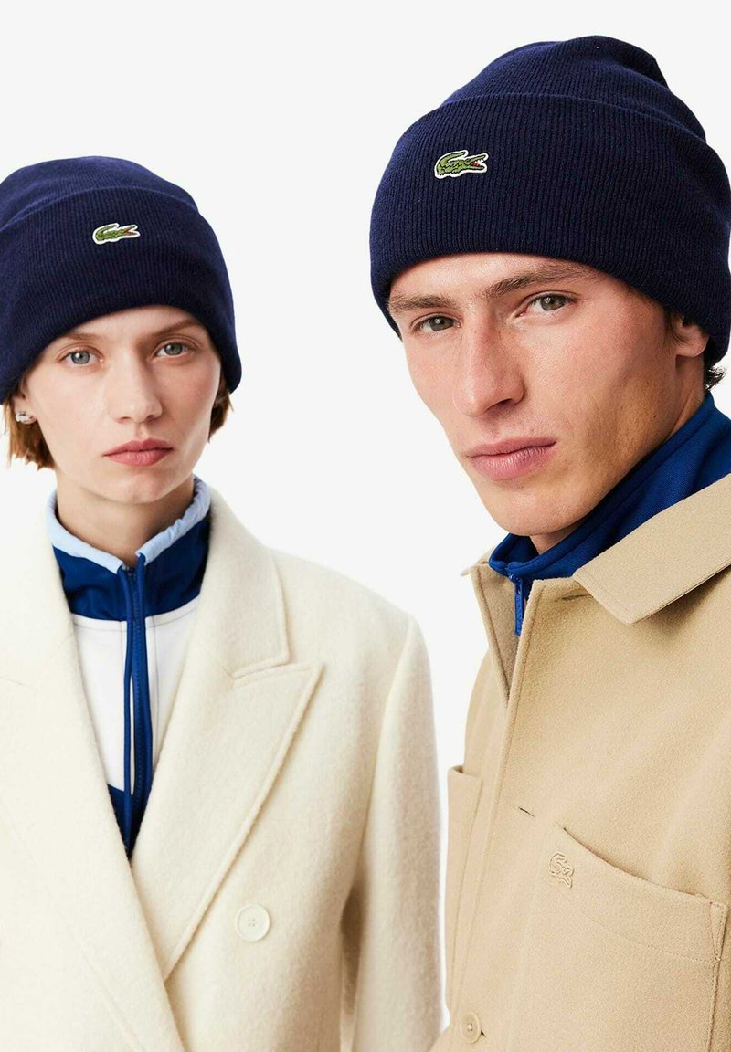 Un bonnet en maille bleu marine foncé avec un logo brodé vert. Mannequins portant des vêtements d'extérieur : un manteau crème à double boutonnage et une veste beige.