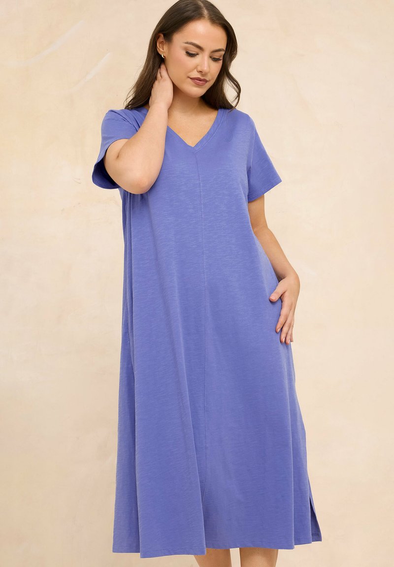 Frau mit langen dunklen Haaren, die ein locker sitzendes, kurzärmeliges Midi-Kleid in Lavendel mit V-Ausschnitt trägt und vor einem schlichten, beigen Hintergrund steht.