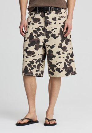 Pantaloncini beige con motivo a macchie di mucca marrone scuro, indossati con cintura nera e infradito nere, mostrati dalla vita ai piedi.