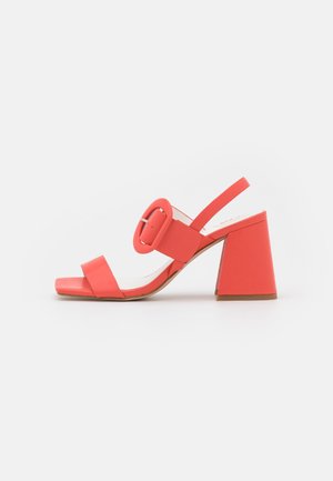 Koralowe sandały na wysokim obcasie z szerokim kwadratowym noskiem, regulowanym paskiem na kostkę, dużym detalem w formie klamry oraz kolorowym, geometrycznym obcasem.