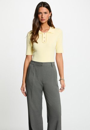 Frau mit langen braunen Haaren, die ein gelbes, kurzärmliges, geripptes Oberteil mit Knöpfen und graue, hochtaillierte, weit geschnittene Hosen trägt, steht.