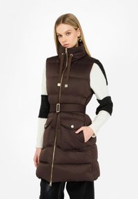 Gilet marrone imbottito con colletto alto, chiusura con zip, vita con cintura e tasche frontali. Presenta maniche texturizzate nere a contrasto.