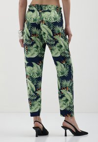 Pantalons à motif floral dans un bleu marine foncé avec des feuilles vertes et des accents orange, dotés d'une taille élastique et de chevilles fuselées, présentés avec des talons noirs.