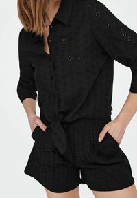 Camicia nera con bottoni davanti e dettaglio con lacci, caratterizzata da motivi floreali traforati. Abbinata a pantaloni shorts neri coordinati, entrambi in un tessuto leggero.