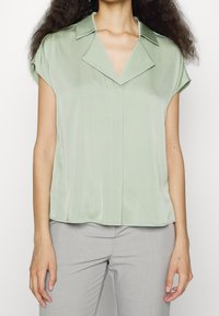 Satinblus i olivgrön med krage och korta ärmar, med knäppning framtill och avslappnad passform. Slät textur som faller mjukt.