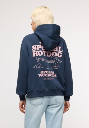 Persona con capelli biondi corti indossa una felpa con cappuccio blu navy con testo rosa e una grafica di un hotdog che corre sulla schiena, abbinata a jeans azzurri.