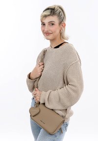 Beige leather crossbody bag with a heart-shaped zipper pull and a smooth texture, worn over a loose-knit beige sweater and light blue jeans.