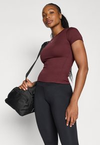 Camiseta ajustada color burdeos de manga corta, hecha de una tela suave, combinada con leggings negros. La modelo sostiene un bolso de gimnasio negro con cremallera.