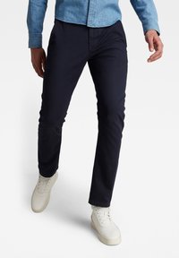 Homme portant un pantalon slim foncé, une chemise en denim bleu clair et des baskets blanches marchant vers l’avant sur un fond blanc.