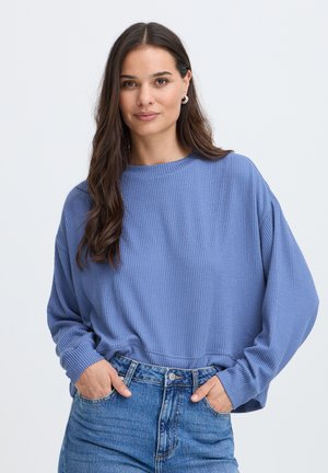 Femme aux longs cheveux bruns portant un pull bleu texturé et un jean bleu, les mains dans les poches, debout devant un fond clair uni.