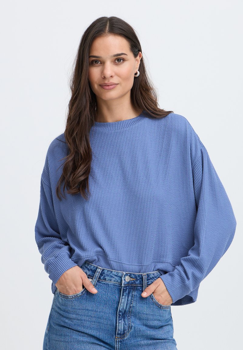 Femme aux longs cheveux bruns portant un pull bleu texturé et un jean bleu, les mains dans les poches, debout devant un fond clair uni.