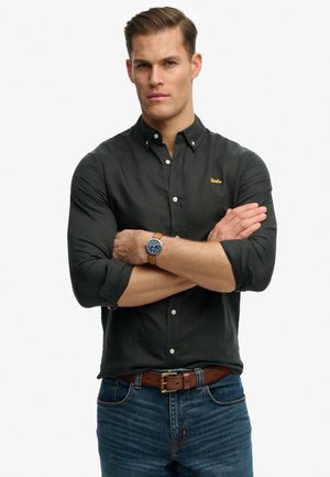 Mann trägt ein schwarzes Hemd mit Knöpfen und blaue Jeans mit verschränkten Armen und zeigt eine braune Lederarmbanduhr am linken Handgelenk.