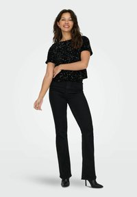 Schwarzes Paillettenshirt mit kurzen Ärmeln, kombiniert mit figurbetonten schwarzen Schlagjeans und schwarzen High-Heels. Glatte Textur, kurzes Design, minimale Details.