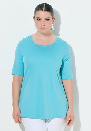 Kobieta w jasnoniebieskiej bluzce z krótkim rękawem ozdobioną kryształkami na dekolcie oraz białych spodniach, stojąca na białym tle.