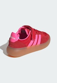 Röd mockaskor med rosa gummidetaljer och tre ränder, rund tå och en robust, genomskinlig gummisula.