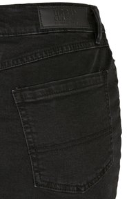 Poche arrière et ceinture d'un jean en denim noir avec des coutures cousues et un patch en cuir noir portant l'étiquette « Urban Classic Denim Factory ».