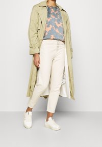 Beige trenchcoat over een blauw en perzikkleurig top met bloemenpatroon, met crèmekleurige broek en witte sneakers, met textuurstof en rechte pasvorm.
